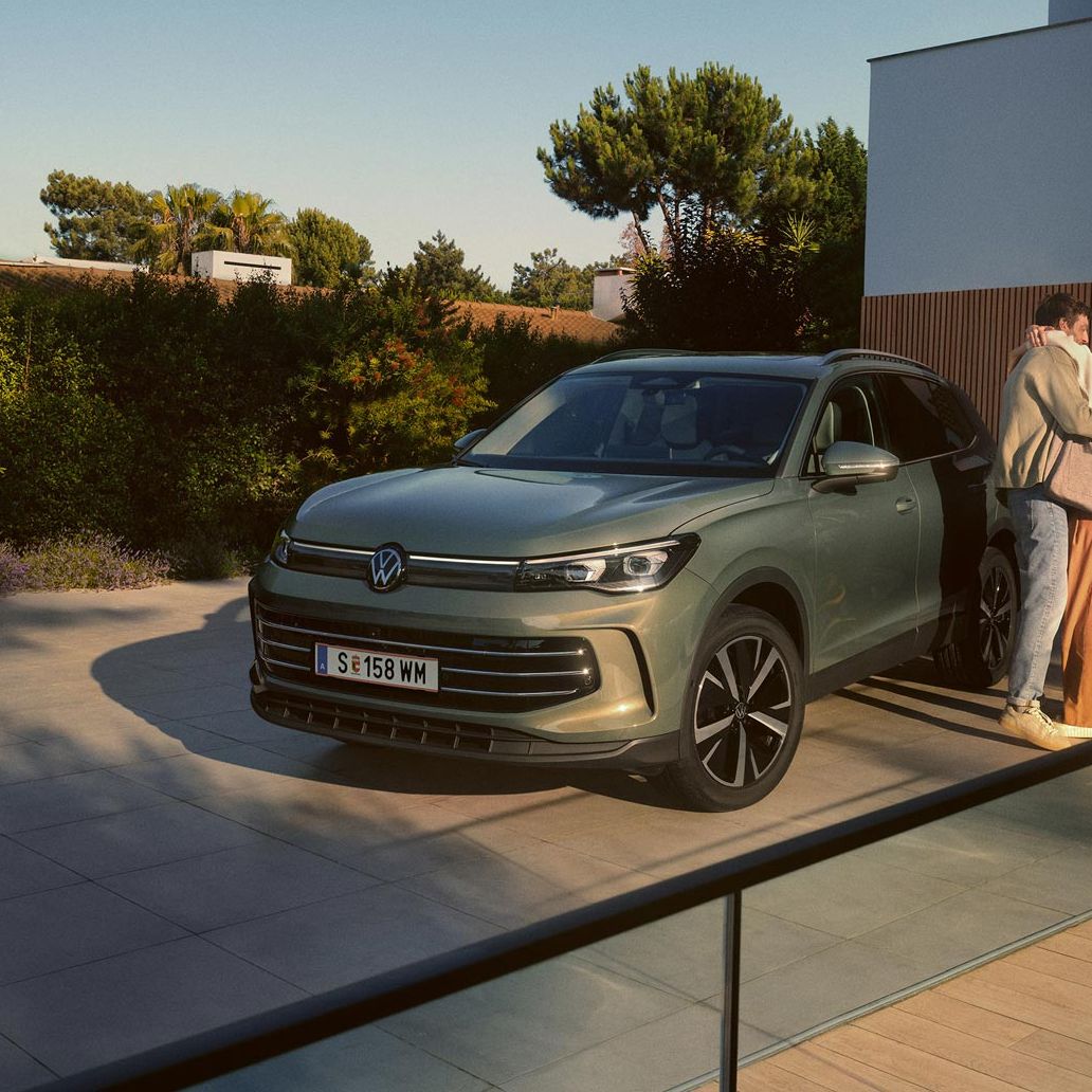 Eine Frau und ein Mann umarmen sich, neben ihnen steht ein VW Tiguan Elegance in der Farbe Cipressino-Grün Metallic