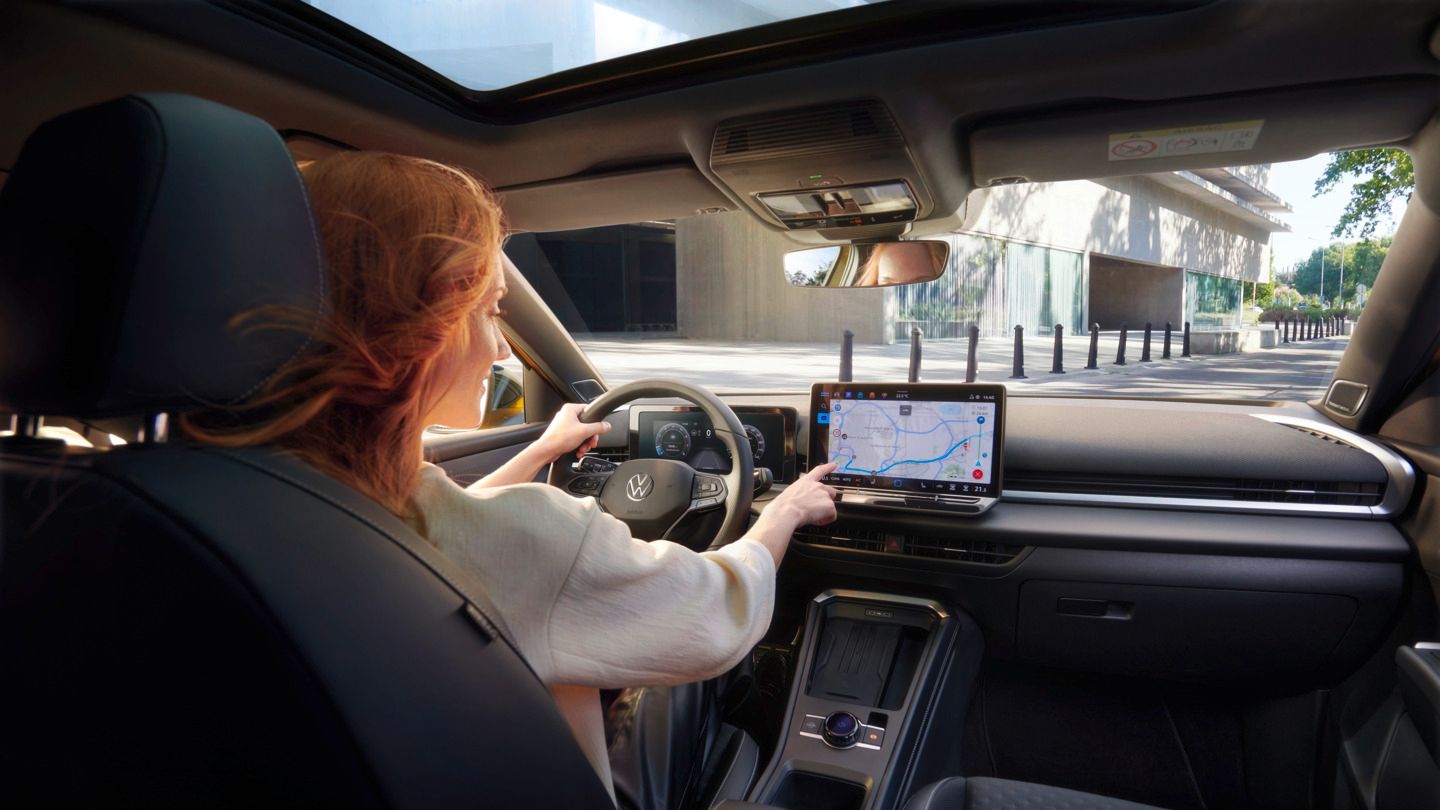 Eine rothaarige Frau sitzt am Fahrersitz eines VW T-Roc Autos. Man sieht die vordere Hälfte des schwarzen Interieurs. Mit der einen Hand bedient sie die Navigation im Infotainmentsystem per Touchscreen, die andere Hand hat sie auf dem Lenkrad. Das Auto scheint durch eine Stadt zu fahren. 