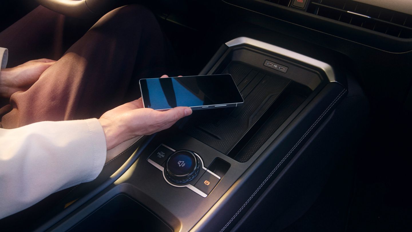 Eine Person hält ein Smartphone und legt es auf die kabellose Ladefläche im Inneren eines Autos. Der Fokus liegt auf der Mittelkonsole des Fahrzeugs. 