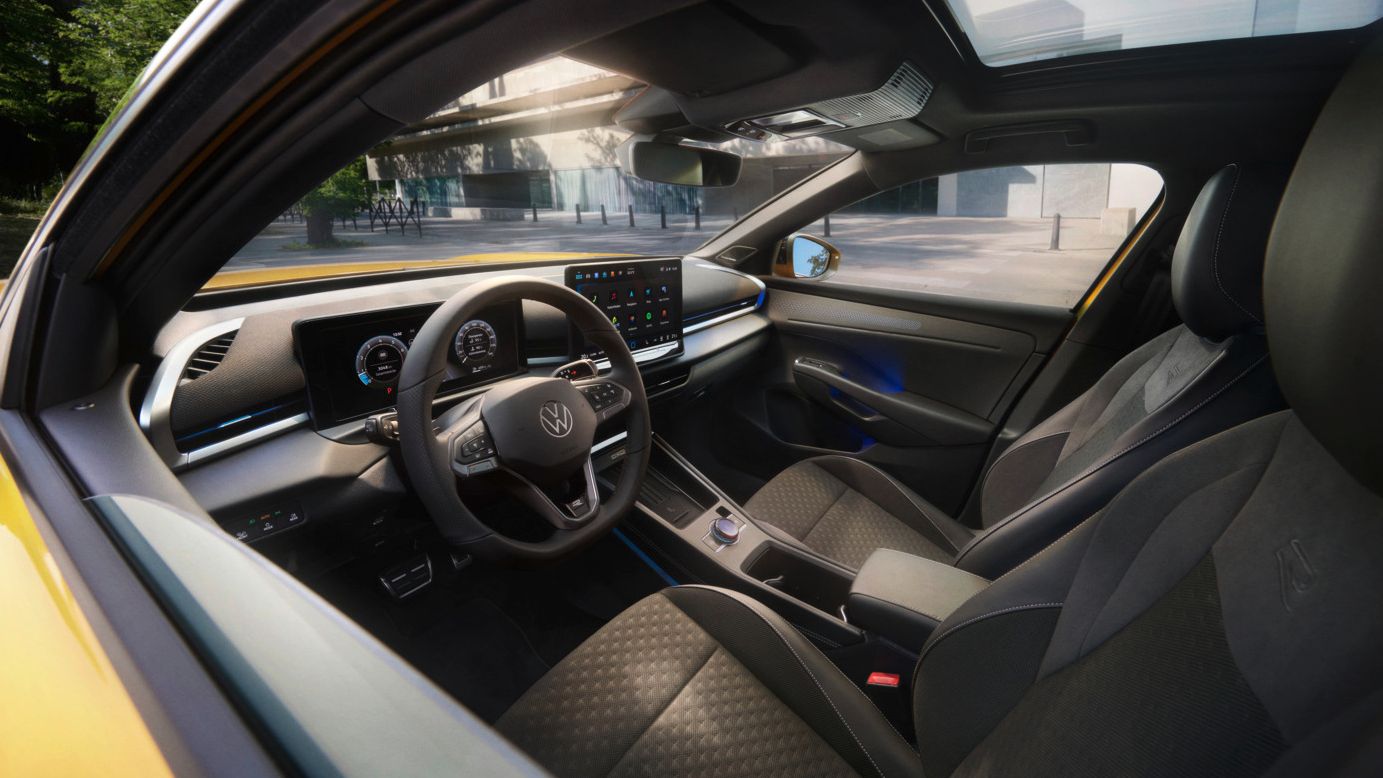 Das bild zeigt den Blick in das Interieur eines gelben VW T-Rocs. Man sieht die beiden vorderen Sitze, ein Lenkrad, das Infotainmentsystem aber auch einen kleinen Teil des gelb lackierten Exterieurs. Im Hintergrund sieht man Ein städtisches Gebäude und daneben dichte Laubbäume.