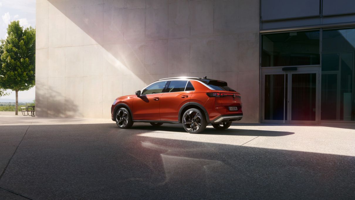 Ein roter T-Roc SUV steht einsam auf einem modernen, weitläufigen Parkplatz neben einem großen Gebäude. Die Sonne scheint und wirft Schatten auf den Boden. Im Hintergrund sind ein Baum und eine Bank zu sehen.