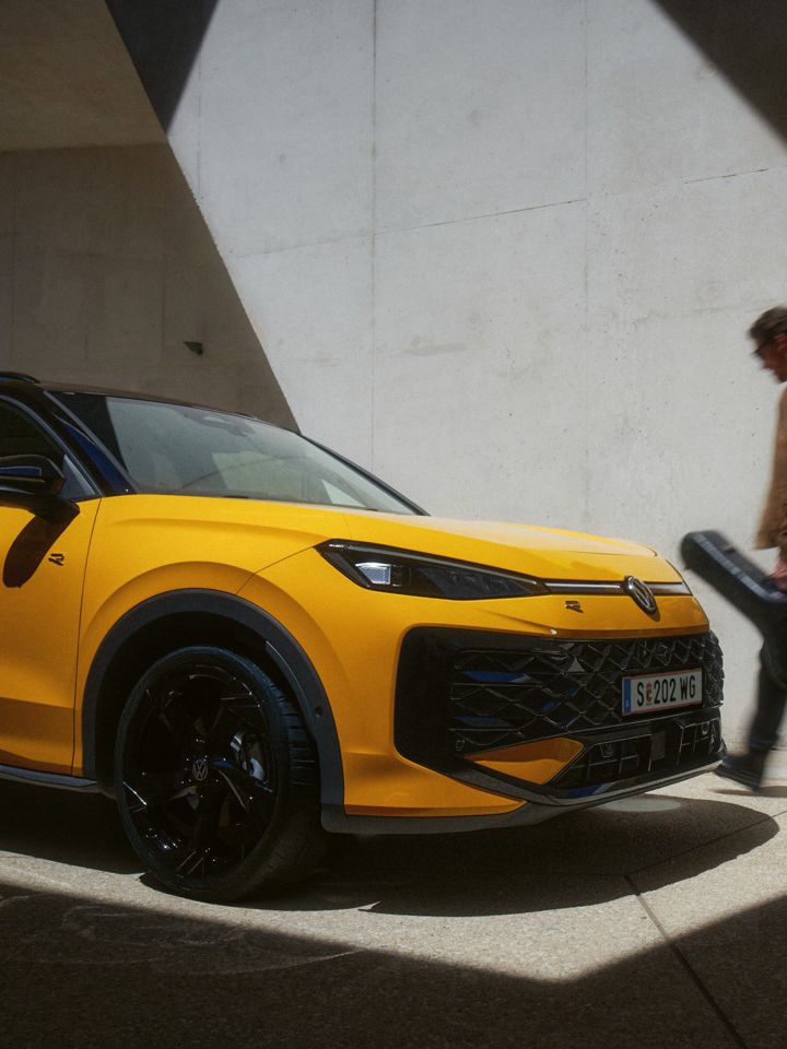 Ein Mann mit Gitarrenkoffer geht an einem gelben VW T-Roc R-Line in moderner Architektur vorbei.