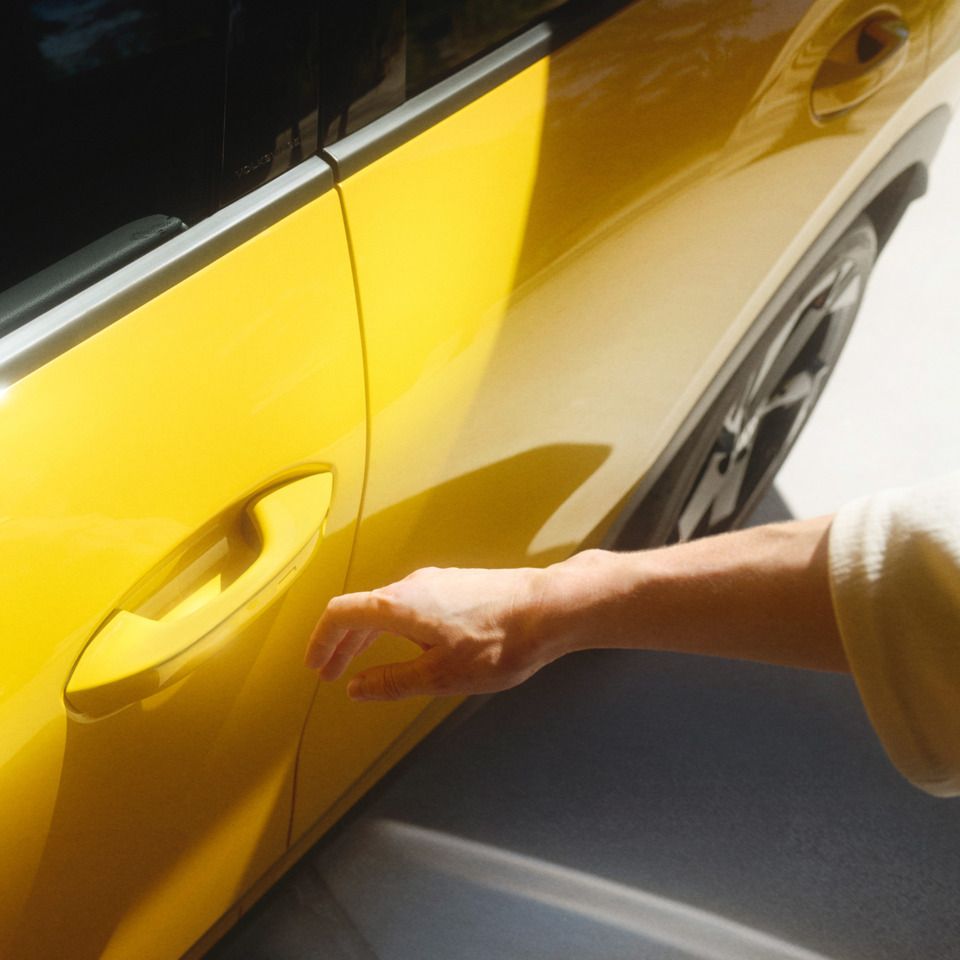 Ein Mensch greift nach dem Türgriff eines gelben Autos. Der Schatten der Hand ist auf dem hellen Boden zu sehen. Das Auto steht still.