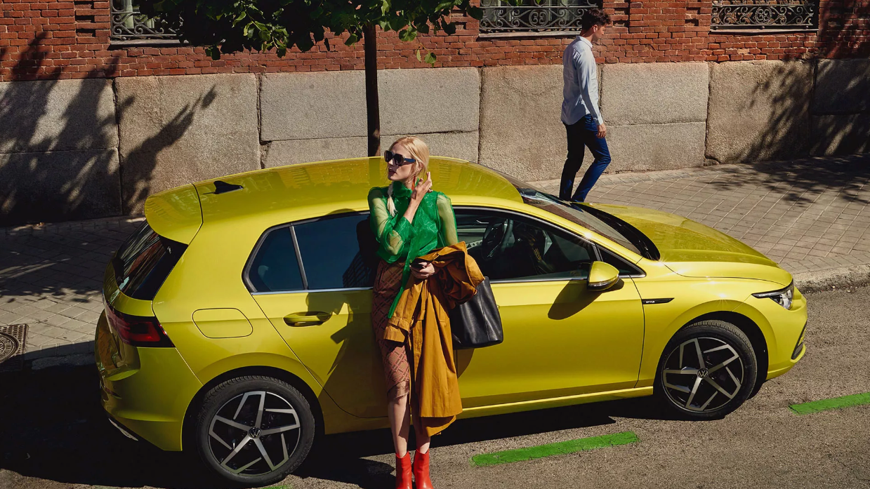 VW Golf i parkuar buzë rrugës, një grua mbështetet në derën anësore.