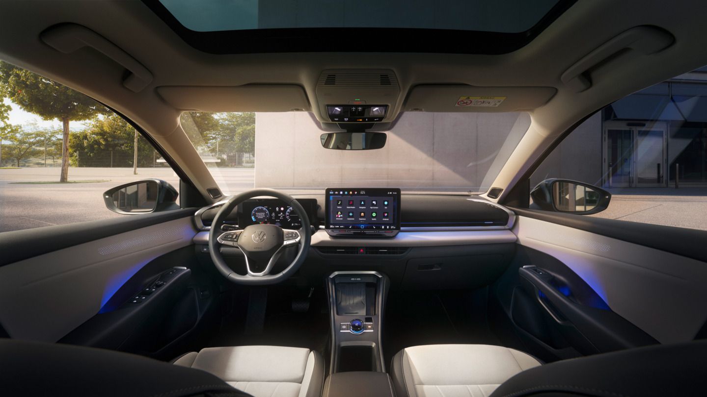 Das Bild zeigt das Innere eines T-Roc mit einem Sonnen- oder Glasdach. Das Cockpit hat ein großes Bildschirm-Display und ein Lenkrad mit mehreren Funktionen. Außen ist eine Grünfläche und ein Gebäude zu erkennen.